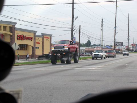 1988 Ford E250 - A heavily modified red pickup truck with a lifted suspension and large tires drives down a suburban street.