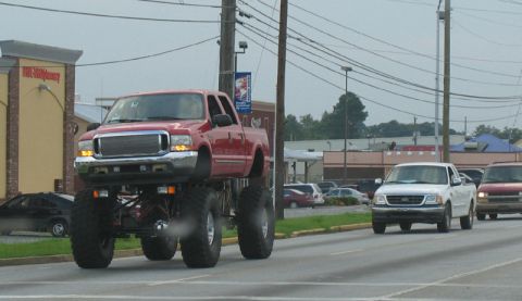 1988 Ford E250 - A red monster truck with massive tires and a lifted suspension drives down the road, passing other vehicles.