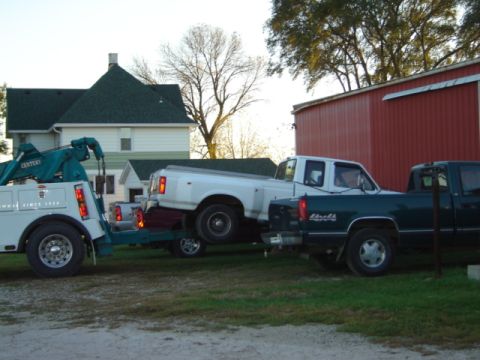 1992 Ford Bronco - A tow truck is hauling two pickup trucks on its trailer in front of a house and a red barn.