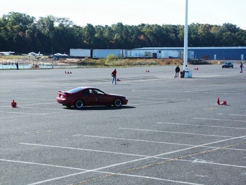 2001 Mercury Grand Marquis - A red coupe navigates a course marked by cones in an open lot, with aircraft and buildings visible in the background.