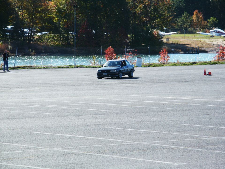Dark sedan on asphalt lot, front 3/4 view, with trees and water in background