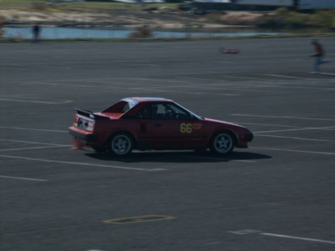 2001 Mercury Grand Marquis - A red sports car with racing numbers is seen on a paved track with a body of water in the background.