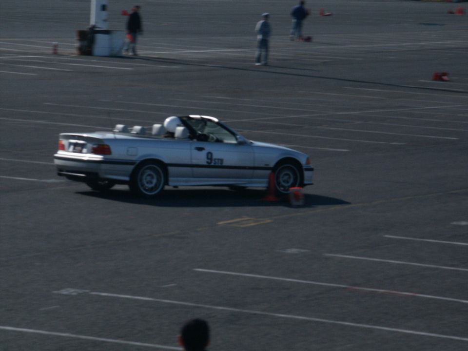 Silver convertible sports car with a white helmet visible in the driver's seat, on a paved track with orange cones