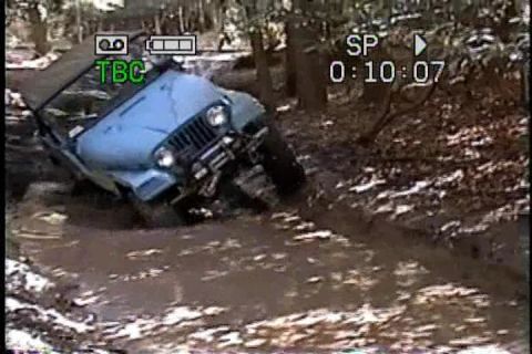 1989 Ford Bronco - A blue Jeep Wrangler navigates a muddy off-road trail, with its front-left wheel deep in the mire.