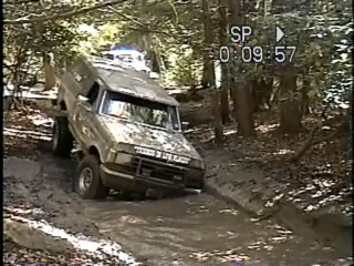 1989 Ford Bronco - An off-road vehicle navigates a challenging muddy terrain surrounded by dense woods.