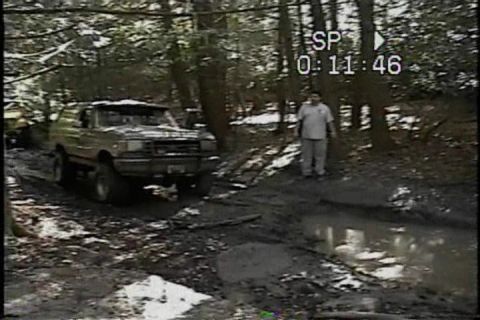 1989 Ford Bronco - A lifted Ford Bronco navigates a muddy off-road course in a wooded area.