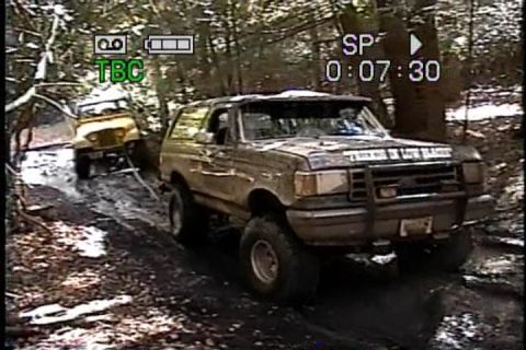 1989 Ford Bronco - Two off-road vehicles navigate a challenging muddy path through a wooded area.