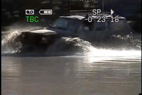 1989 Ford Bronco - A dark-colored SUV drives through a body of water, creating a large splash.