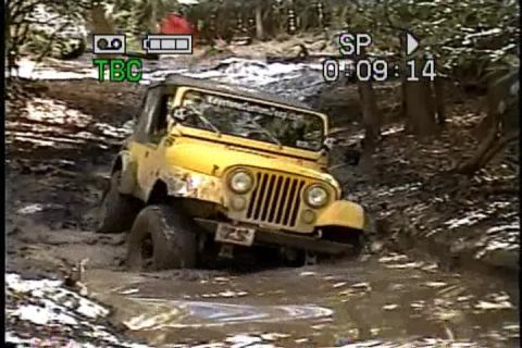 1989 Ford Bronco - A yellow Jeep Wrangler navigates a muddy trail with trees on either side.