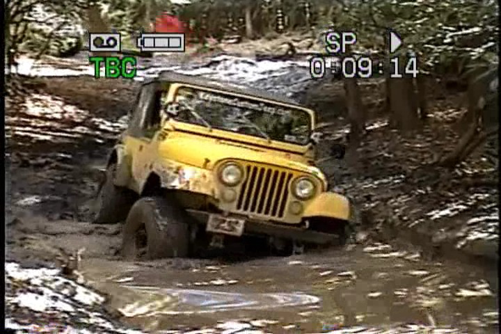 Yellow Jeep Wrangler driving through mud, front grille visible, windshield cracked