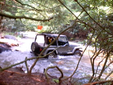 2001 Ford Escape - A gray Jeep Wrangler navigates a rocky stream surrounded by dense foliage.