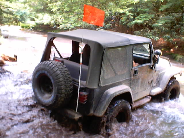 Muddy soft-top Jeep Wrangler with orange trail flag fording a rocky creek