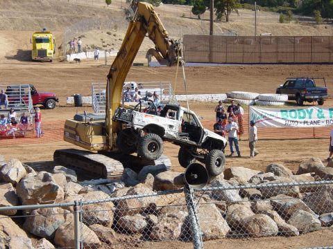 1986 Ford Bronco - An excavator lifts a modified off-road truck during an event, with spectators and other vehicles present.