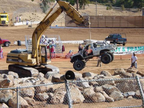 1986 Ford Bronco - An excavator lifts a modified white truck during an event.