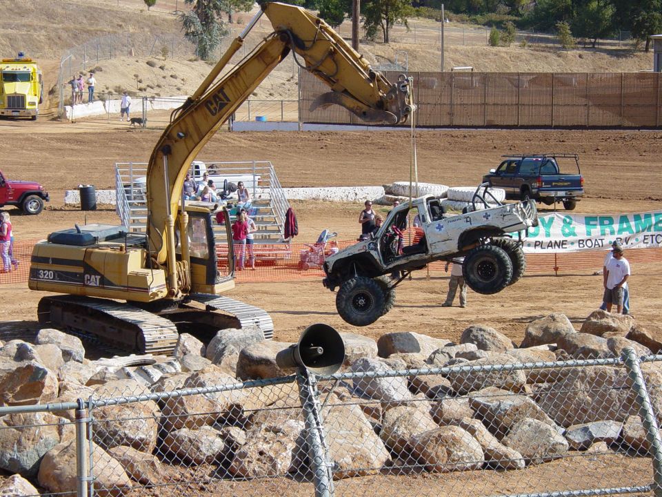 White lifted truck with roll cage suspended by excavator arm over rocks