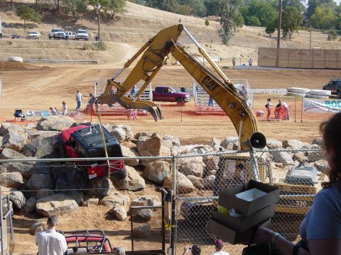 1986 Ford Bronco - An excavator is lifting a wrecked Jeep over a rocky area at an outdoor event.