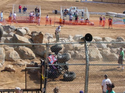 1986 Ford Bronco - A monster truck has overturned during an event, with onlookers observing from the stands and trackside.