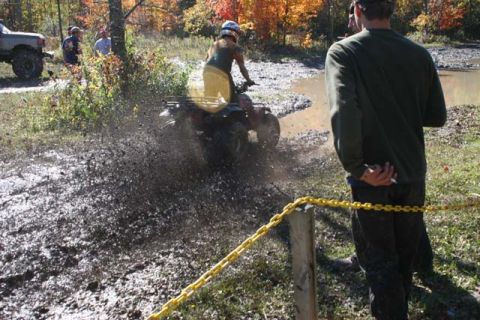 1987 Ford Bronco - An ATV kicks up a large spray of mud while navigating an off-road course, with onlookers observing the action.