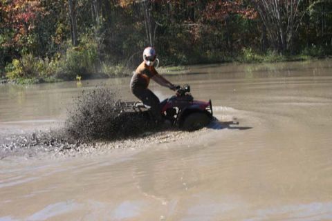 1987 Ford Bronco - Rider on a red ATV kicks up a large spray of mud as they navigate through a shallow body of water.