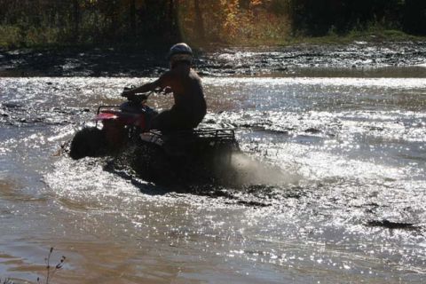 1987 Ford Bronco - A person rides an all-terrain vehicle through a muddy, watery area, kicking up spray.