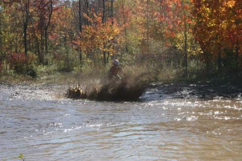 1987 Ford Bronco - An ATV rider creates a large splash as they navigate through a muddy, watery trail surrounded by autumn trees.