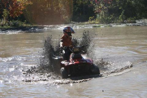 1987 Ford Bronco - A rider on a red ATV navigates through a muddy water obstacle, sending up a large spray.