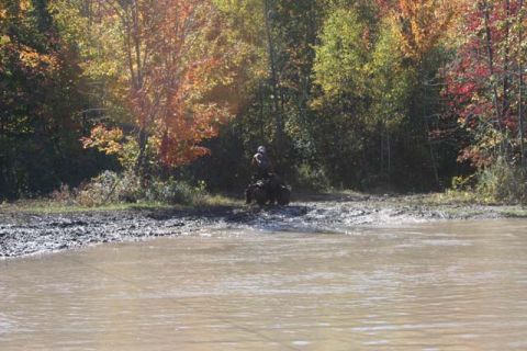 1987 Ford Bronco - An all-terrain vehicle navigates a muddy area next to a body of water, surrounded by trees in fall colors.