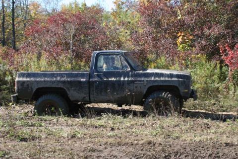 1987 Ford Bronco - A black pickup truck with off-road tires is stuck in muddy terrain surrounded by autumn foliage.