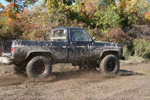 1987 Ford Bronco - Muddy black pickup truck navigates a wet, off-road course with trees in the background.