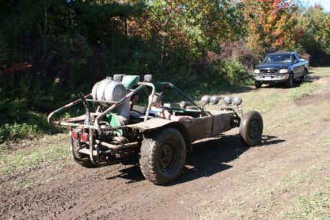 1987 Ford Bronco - A muddy dune buggy navigates a dirt path, with a pickup truck visible in the background.