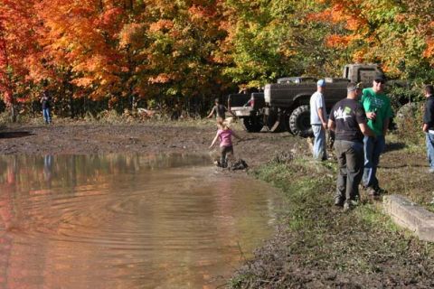 1987 Ford Bronco - People and trucks are gathered in a muddy area with colorful autumn trees in the background.