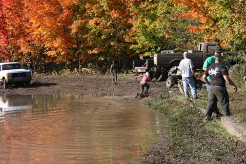 1987 Ford Bronco - Several vehicles are engaged in off-roading activities near a body of water, surrounded by trees with colorful autumn leaves.