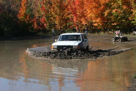 1987 Ford Bronco - A white 1987 Ford Bronco navigates a muddy off-road course during autumn, with an ATV visible in the background.