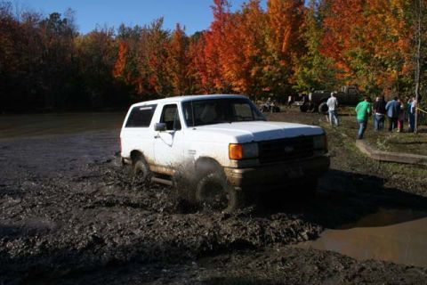 1987 Ford Bronco - A white SUV navigates a muddy trail with onlookers and other vehicles nearby during autumn.