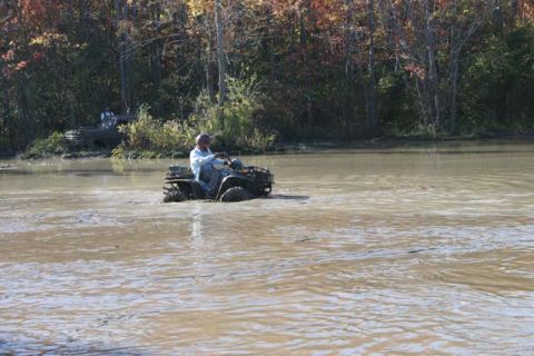 1987 Ford Bronco - ATV rider navigates through muddy water with another vehicle visible in the distance.