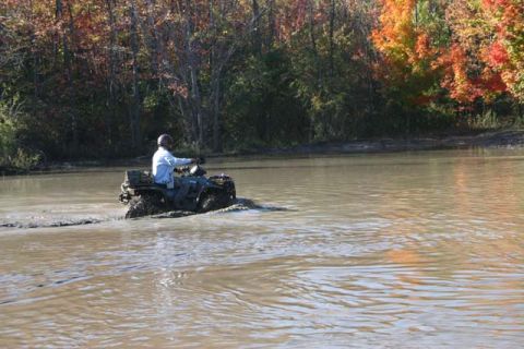 1987 Ford Bronco - A person rides an all-terrain vehicle through a muddy body of water with colorful autumn trees in the background.