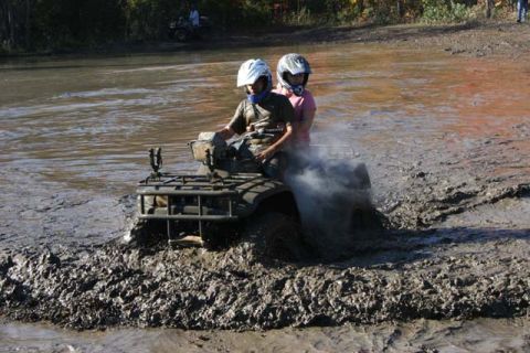 1987 Ford Bronco - Two individuals ride an ATV through a muddy terrain, kicking up spray.
