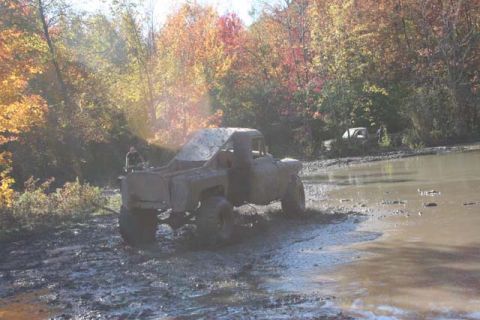 1987 Ford Bronco - A muddy pickup truck navigates a challenging off-road terrain with autumn foliage surrounding the area.