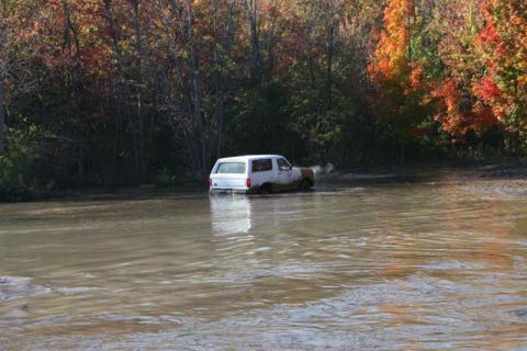 1987 Ford Bronco - A white SUV is partially submerged in muddy water, with trees displaying autumn foliage in the background.