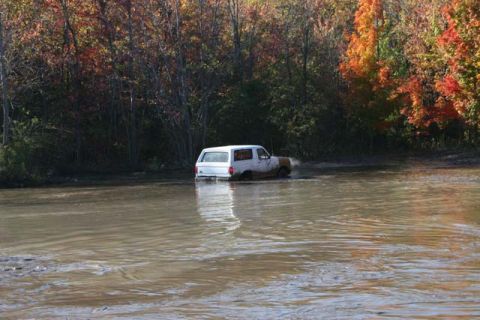 1987 Ford Bronco - A white SUV navigates through a muddy body of water surrounded by trees displaying fall colors.
