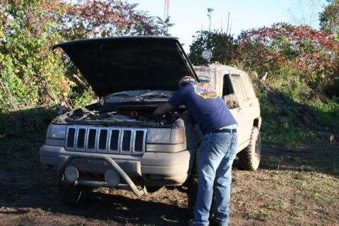 1987 Ford Bronco - A person works on the engine of a mud-covered Jeep Grand Cherokee parked in an outdoor, wooded area.