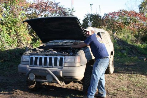 1987 Ford Bronco - A man inspects the engine of a dirty SUV parked on a dirt road with trees in the background.