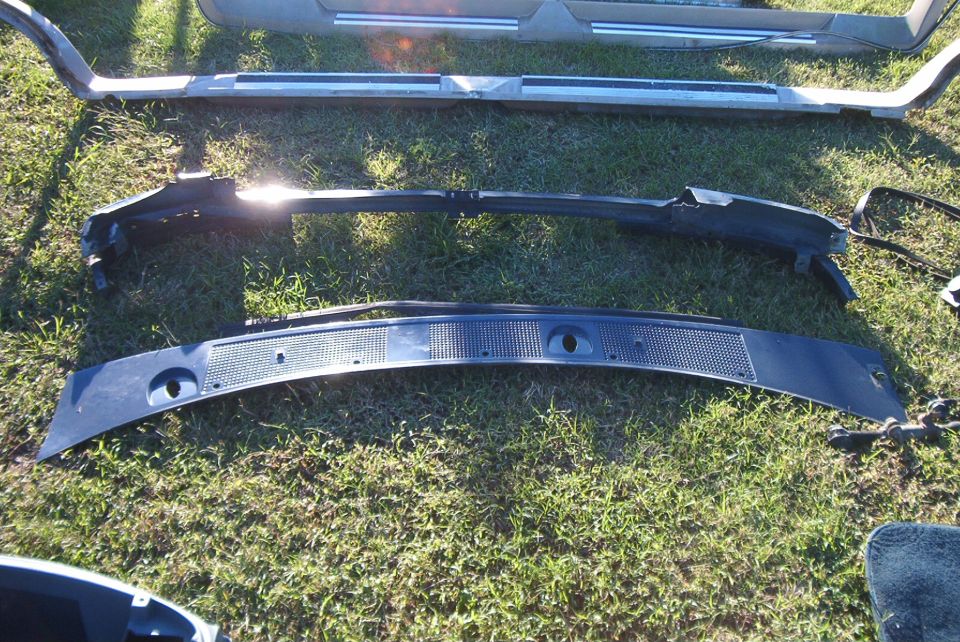 Several car parts, including a black plastic cowl vent panel and a chrome trim piece, laid out on grass