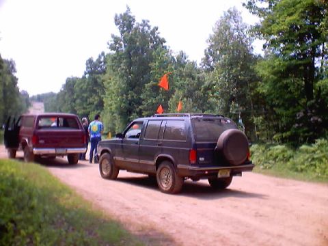 2001 Ford Escape - A dark SUV with a spare tire on the back is parked on a dirt road next to a maroon pickup truck.