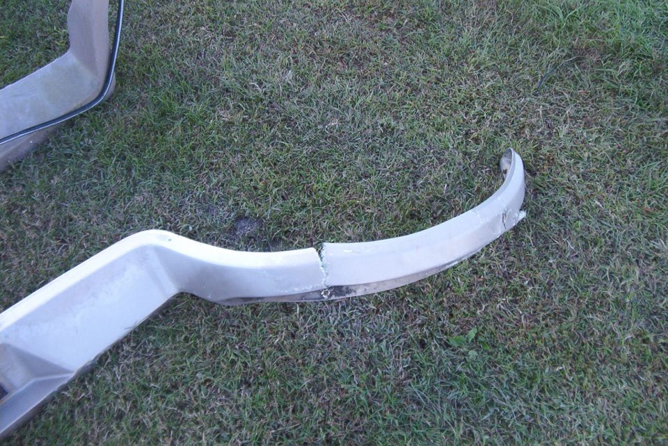 Broken white bumper section on grass. The part is cracked in the middle and shows damage on the right end
