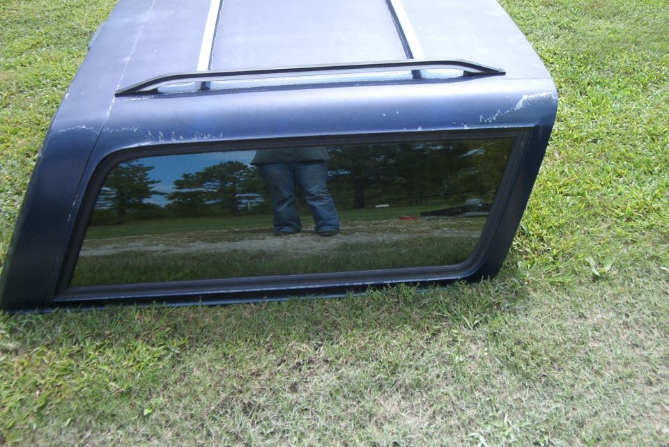 Dark blue truck cap with a window and roof rack, sitting on grass
