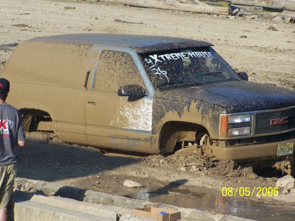 Mud-covered GMC Suburban stuck in mud, front grille and headlights visible, '4XTREMEHBUSE' sticker on windshield