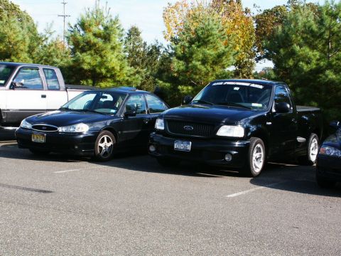 2001 Mercury Grand Marquis - A black pickup truck is parked next to a sedan, with trees in the background.