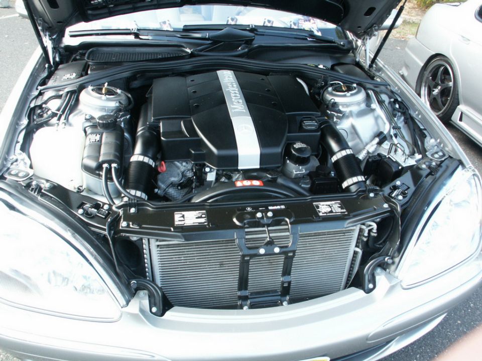Silver Mercedes-Benz engine bay with "Mercedes-Benz" lettering on the engine cover. Another car is visible to the right