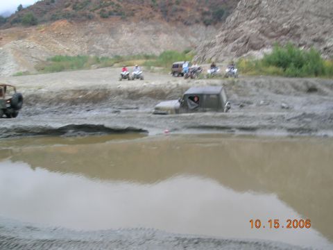 1985 Ford Bronco - Multiple off-road vehicles navigate a muddy terrain, with one vehicle partially submerged in deep mud.